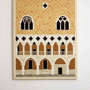 May include: A quilted wall hanging depicting a building with a patterned facade, arched windows, and a balcony. The building is rendered in shades of orange, white, and brown.