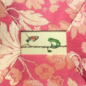 May include: A framed painting of a green tree frog sitting on a branch with a butterfly flying towards it. The painting is set against a pink floral background.