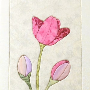 Può includere: Un'opera d'arte in tessuto incorniciata che presenta un design stilizzato di tre tulipani rosa e viola con steli e foglie verdi.