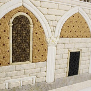 May include: A close-up of a white brick wall with two arched doorways and a window. The doorways have a brown and gold patterned fabric behind them. The window has a black and brown patterned glass.