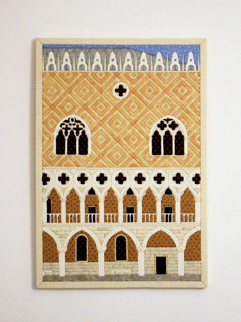 May include: A quilted wall hanging depicting a building with a patterned facade, arched windows, and a balcony. The building is rendered in shades of orange, white, and brown.