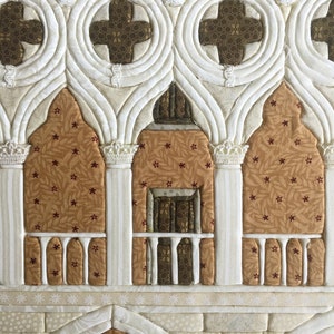 May include: A white fabric quilt with a brown and white pattern depicting a building with arched windows and a balcony. The quilt is made with a variety of fabrics and textures.