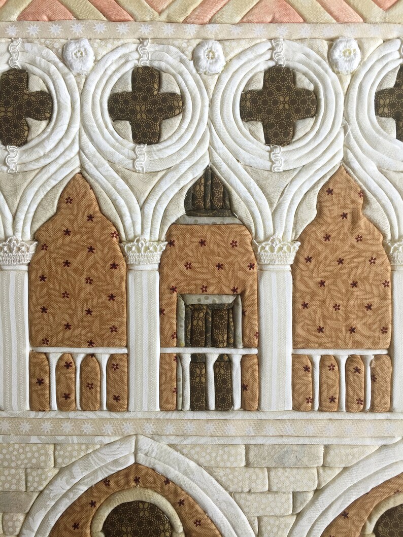 May include: A white fabric quilt with a brown and white pattern depicting a building with arched windows and a balcony. The quilt is made with a variety of fabrics and textures.