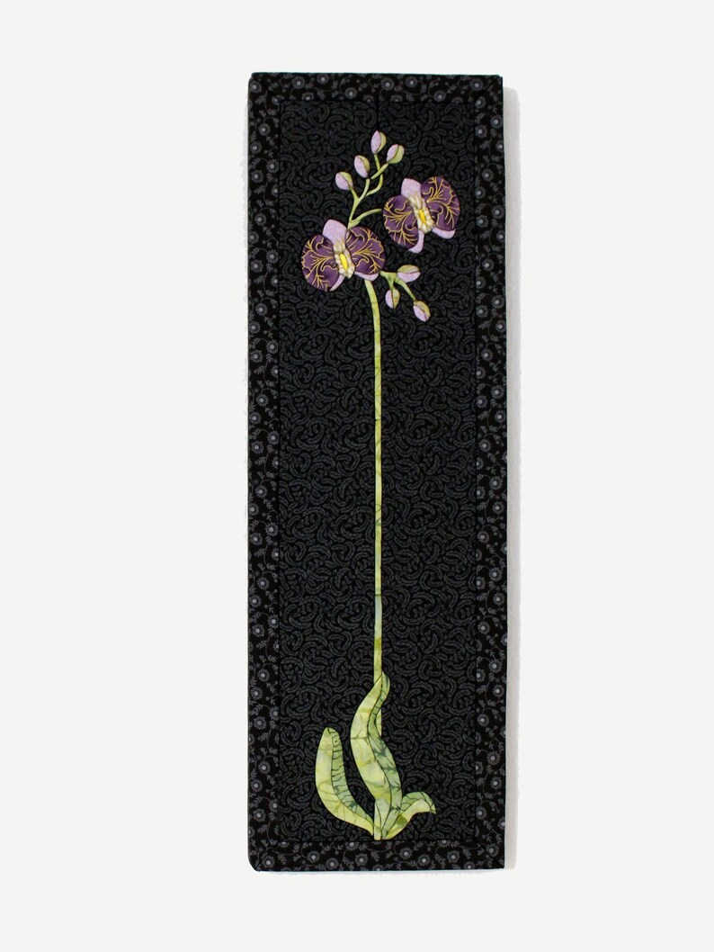 May include: A black framed wall art piece with a single purple orchid flower with green leaves on a black background.