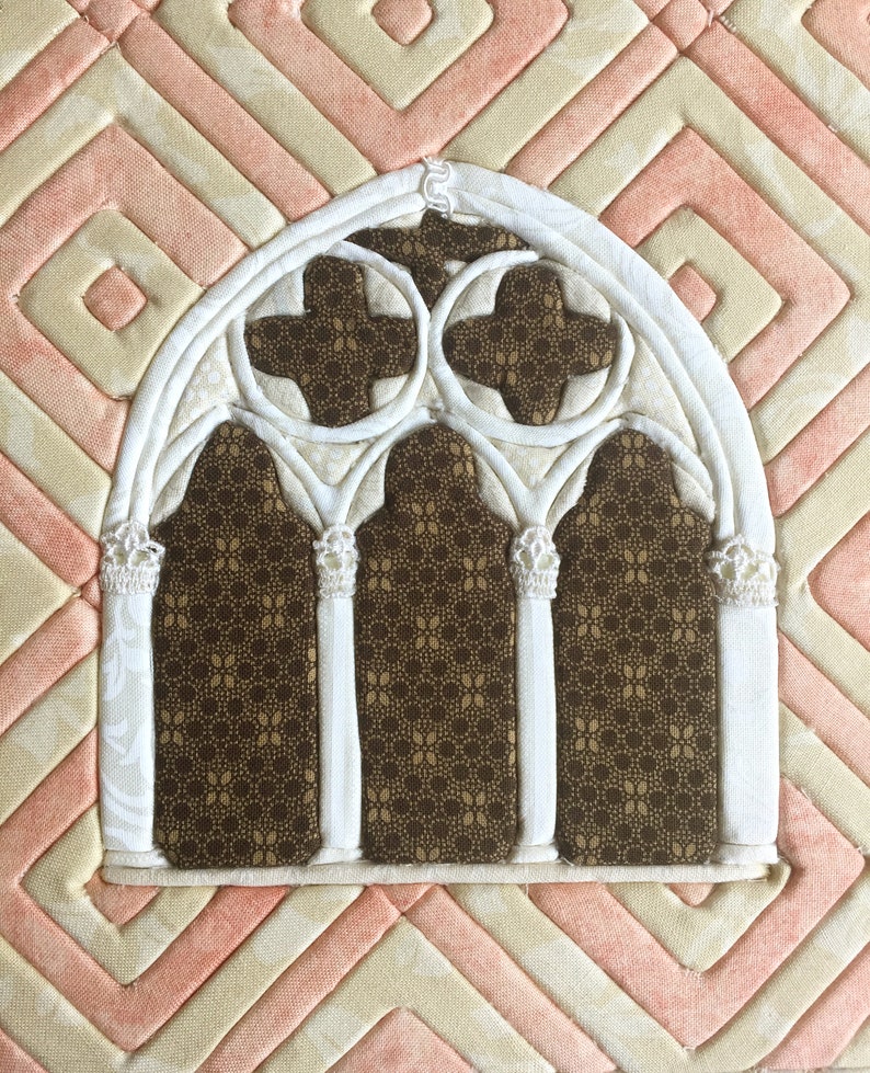 May include: A white fabric applique of a gothic arch window with three arched sections and a brown fabric background with a repeating pattern.