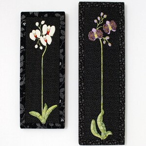 May include: Two framed black and white fabric art pieces featuring embroidered orchid flowers. The orchid on the left is white with red accents, and the orchid on the right is purple with yellow accents.