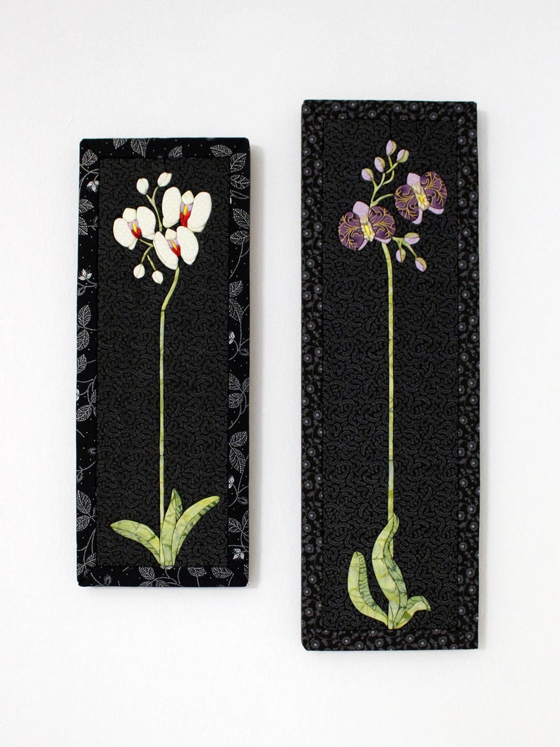 May include: Two framed black and white fabric art pieces featuring embroidered orchid flowers. The orchid on the left is white with red accents, and the orchid on the right is purple with yellow accents.