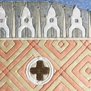 May include: A quilted wall hanging with a peach and cream geometric pattern. The centre of the piece features a brown cross surrounded by a white circle.
