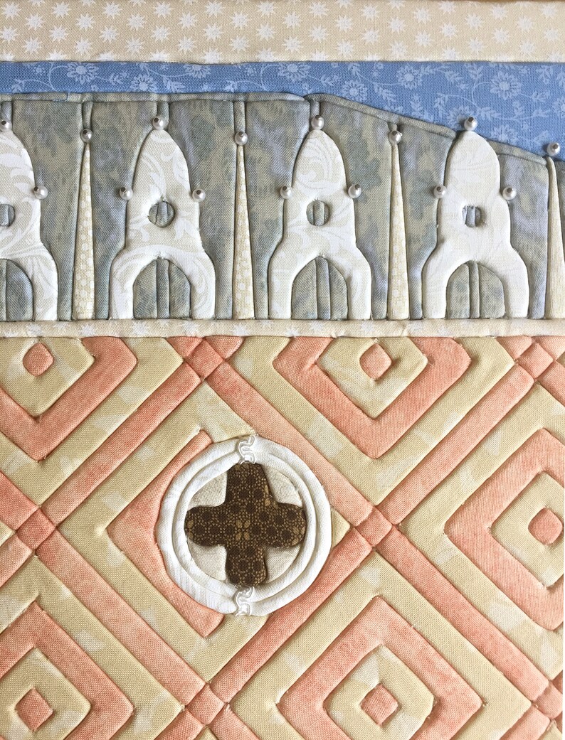May include: A quilted wall hanging with a peach and cream geometric pattern. The centre of the piece features a brown cross surrounded by a white circle.