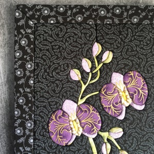 May include: A close-up of a purple and gold embroidered orchid flower on a black fabric background with a white floral pattern.