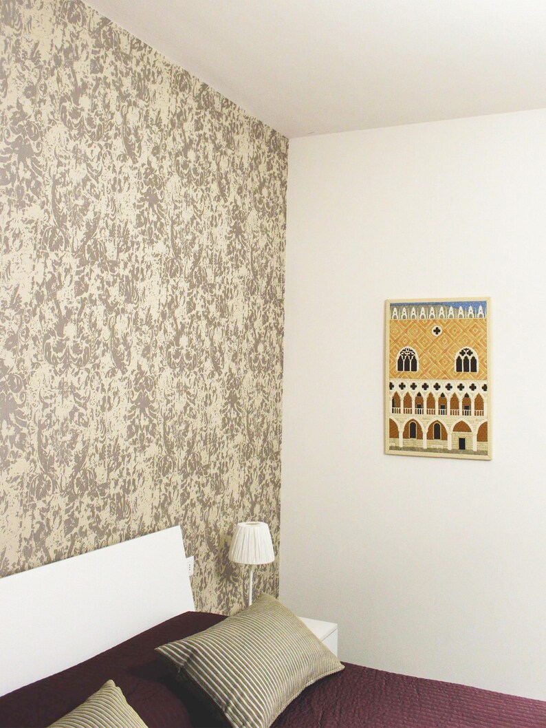 May include: A white bed with a burgundy bedspread and two striped pillows. A white lamp sits on a bedside table next to the bed. A framed print of a building with arches hangs on the wall behind the bed.