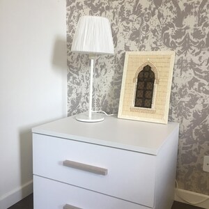 May include: White nightstand with two drawers and a white lamp with a white shade. The nightstand is against a wall with a beige and white patterned wallpaper. A framed picture of a mosaic archway is on top of the nightstand.