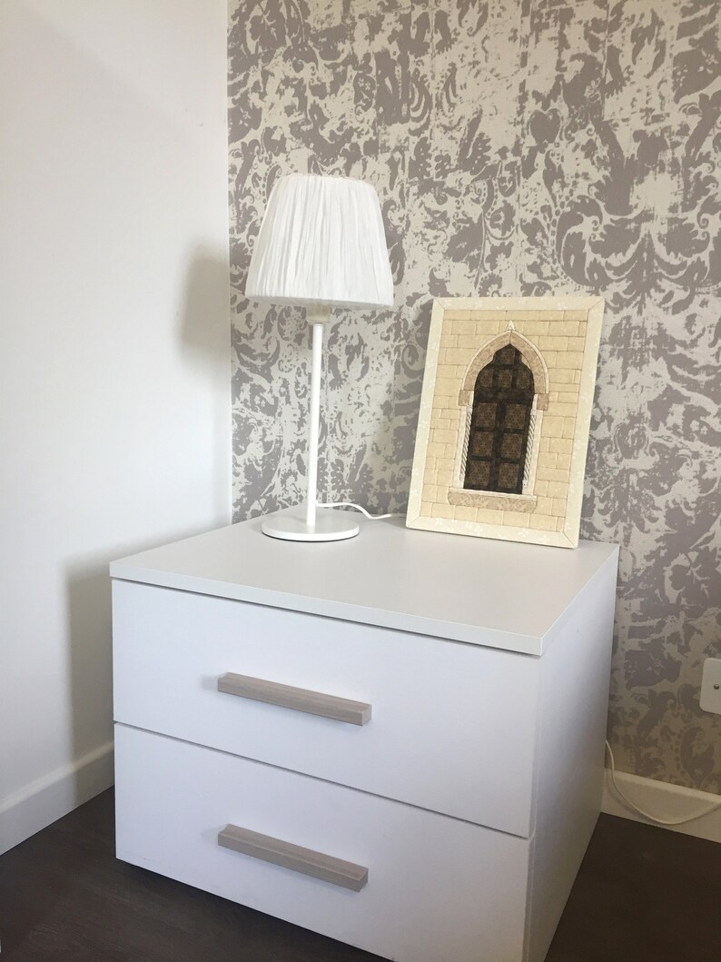 May include: White nightstand with two drawers and a white lamp with a white shade. The nightstand is against a wall with a beige and white patterned wallpaper. A framed picture of a mosaic archway is on top of the nightstand.
