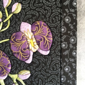 May include: Close-up of a purple and gold fabric orchid flower with a white center, on a black background with a floral pattern.