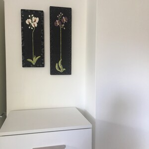 May include: Two framed black and white floral prints with white orchids and purple orchids on a white wall above a white dresser with two drawers.