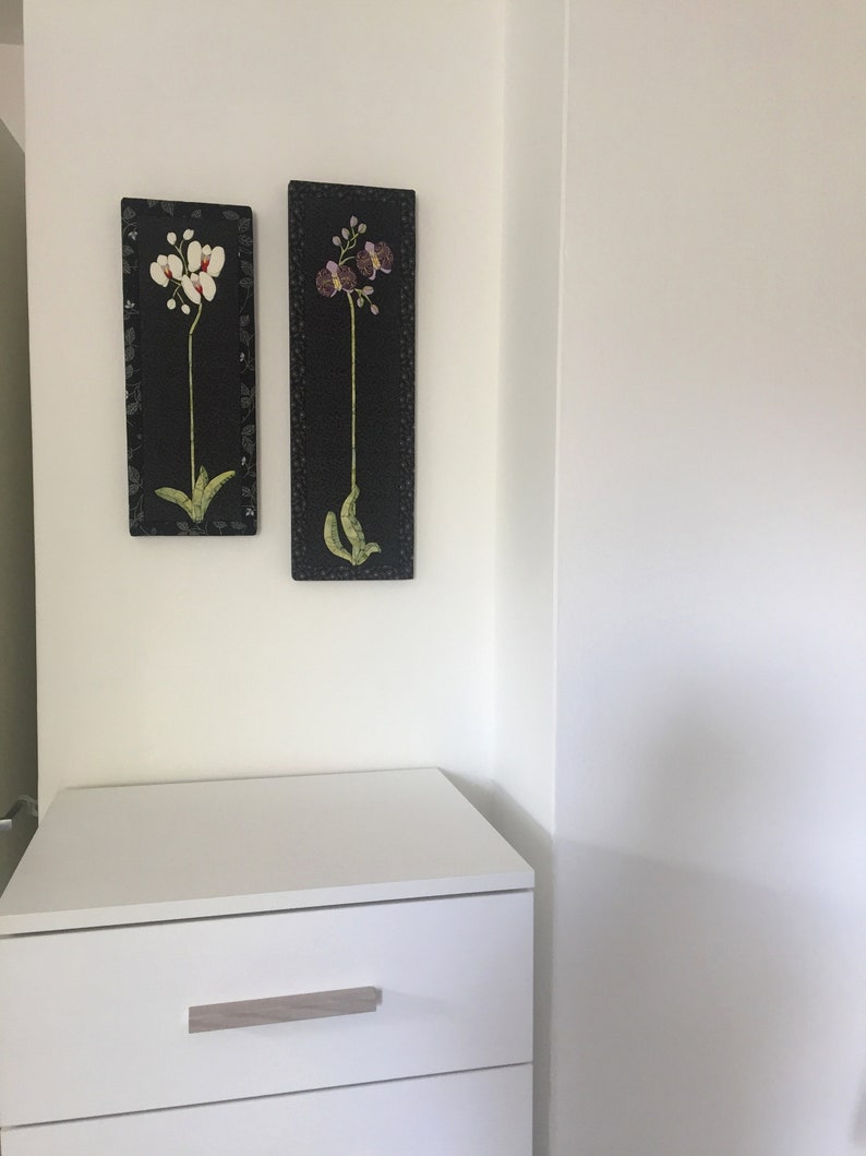 May include: Two framed black and white floral prints with white orchids and purple orchids on a white wall above a white dresser with two drawers.