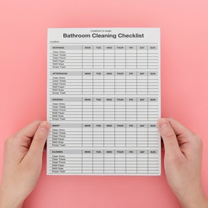 May include: A white bathroom cleaning checklist with a gray grid, held by two hands against a pink background. The checklist is divided into morning, afternoon, evening, night, and closing sections, with columns for each day of the week.