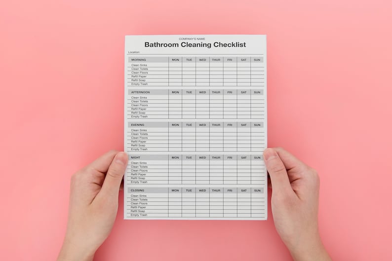 May include: A white bathroom cleaning checklist with a gray grid, held by two hands against a pink background. The checklist is divided into morning, afternoon, evening, night, and closing sections, with columns for each day of the week.