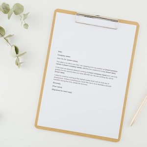 May include: A clipboard with a white sheet of paper and a silver clip, with a resignation letter printed on it. A gold pen rests beside the clipboard. Eucalyptus leaves and a small green leaf are arranged around the clipboard on a white surface.