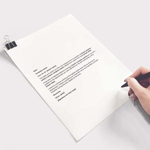 May include: A white sheet of paper with printed text, held in place by a black binder clip. A hand holding a black pen with a red tip is writing on the paper. The paper is angled, with a plain white background.