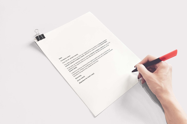 May include: A white sheet of paper with printed text, held in place by a black binder clip. A hand holding a black pen with a red tip is writing on the paper. The paper is angled, with a plain white background.