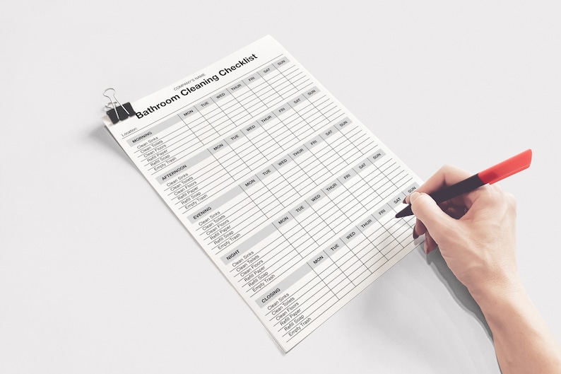 May include: A bathroom cleaning checklist with a black binder clip. The checklist is being filled out with a red and black pen. The checklist includes sections for morning, afternoon, evening, night, and closing.