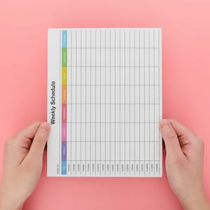 May include: A white weekly schedule planner with a colorful vertical column on the left side. The planner has a grid layout with lines for each day of the week and time slots. The planner is being held by two hands.
