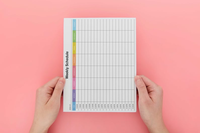 May include: A white weekly schedule planner with a colorful vertical column on the left side. The planner has a grid layout with lines for each day of the week and time slots. The planner is being held by two hands.