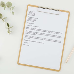 Może przedstawiać: List z rezygnacji na tablicy z klipsem i złotym długopisem. List jest napisany na białym papierze z brązową ramką. List zawiera temat "List z rezygnacji".