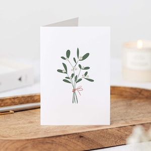 May include: A folded white greeting card featuring a green and white mistletoe illustration tied with a red bow. The card is on a wooden tray with a pen and a candle in the background. The card is a festive holiday item.