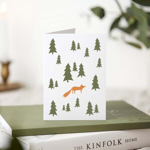 May include: A folded greeting card with a forest scene. The white card features a repeating pattern of green pine trees and an orange fox. The card is resting on a stack of books with a green cover.