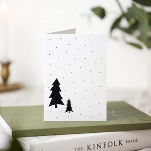 May include: A white greeting card with a black tree design and small black dots. The card is folded and rests on a stack of books. The overall theme is a minimalist holiday or winter design.
