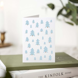 May include: A white greeting card featuring a pattern of light blue Christmas trees with brown trunks and white dots. The card is propped up on a stack of books, with a lit candle and greenery in the background.