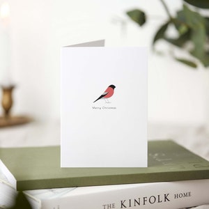 May include: A folded white Christmas card featuring a minimalist illustration of a red and black bird. The words "Merry Christmas" are printed below the bird. The card is resting on a stack of books with a lit candle in the background.