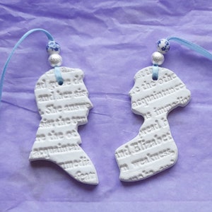 Puede incluir: Dos adornos de cerámica blanca con forma de siluetas, cada uno con texto. Están suspendidos por cintas azules y adornados con cuentas decorativas. El fondo es una superficie texturizada de color lila claro.