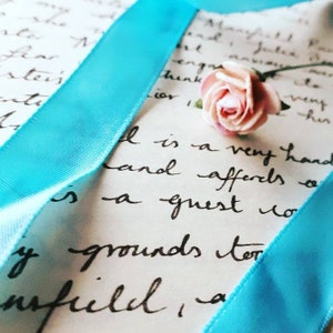 May include: A handwritten letter with a light blue ribbon and a pink rose on top. The letter is written in cursive and the ribbon is tied in a bow.