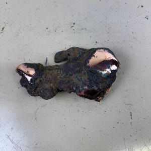 May include: A weathered piece of copper with a dark, mottled surface and patches of bright, polished copper. The irregular shape suggests it may be a fragment of a larger object or a raw piece of the metal. The copper has a mix of green and brown oxidation.