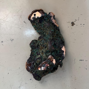 May include: A rough, irregularly shaped piece of copper ore with a dark, textured surface. Patches of green and copper-colored oxidation are visible, creating a contrast against the darker areas. The ore appears to be raw and unprocessed.