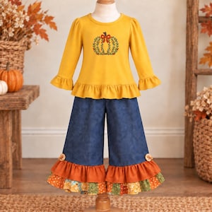 May include: A mustard yellow long-sleeve top with a ruffled hem and a pumpkin embroidery on the chest, paired with dark blue denim-style pants featuring a multi-layered ruffled hem in orange and a floral pattern with small wooden buttons.