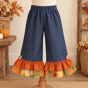 May include: Dark blue denim pants featuring an elastic waistband and wide legs. Each leg hem is finished with a solid burnt orange ruffle layer followed by a multi-colored patchwork ruffle layer with floral and polka dot patterns and wooden buttons.