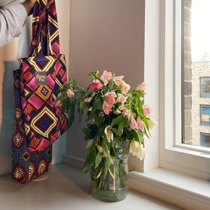 Pode incluir: Uma bolsa tote rosa e azul com um padr&atilde;o geom&eacute;trico e o texto "Bijou Bazaar" pendurada em uma parede branca. Um vaso de flores rosa est&aacute; em um balc&atilde;o branco ao lado de uma janela.
