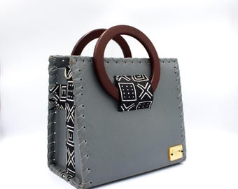 Puede incluir: Un bolso de mano de cuero gris con asa de madera y una correa con estampado en blanco y negro. El bolso tiene un diseño cosido y un logotipo dorado.