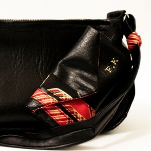 May include: Black leather shoulder bag with a red and gold patterned scarf tucked inside. The bag has a zip closure and a gold metal buckle. The scarf has the initials "F.K." embroidered on it.