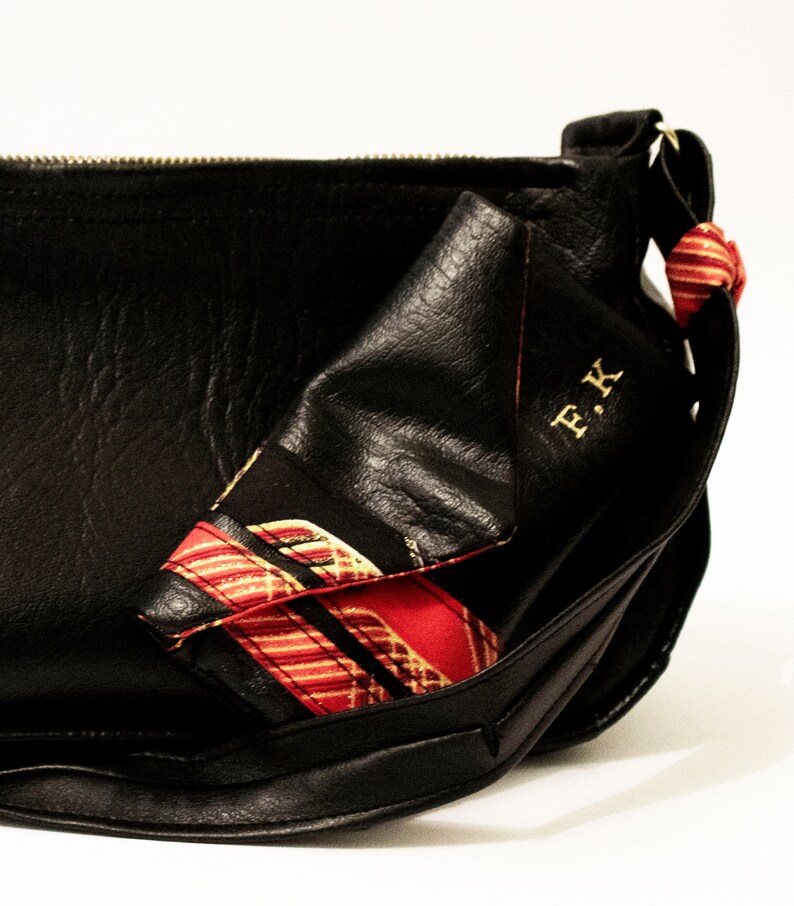 May include: Black leather shoulder bag with a red and gold patterned scarf tucked inside. The bag has a zip closure and a gold metal buckle. The scarf has the initials "F.K." embroidered on it.