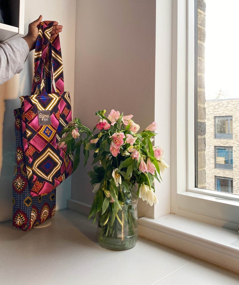 Pode incluir: Uma bolsa tote rosa e azul com um padr&atilde;o geom&eacute;trico e o texto "Bijou Bazaar" pendurada em uma parede branca. Um vaso de flores rosa est&aacute; em um balc&atilde;o branco ao lado de uma janela.