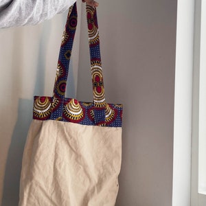 Pode incluir: Uma bolsa tote de lona bege com uma borda de tecido estampado em azul, vermelho e dourado. A bolsa tem duas al&ccedil;as longas.