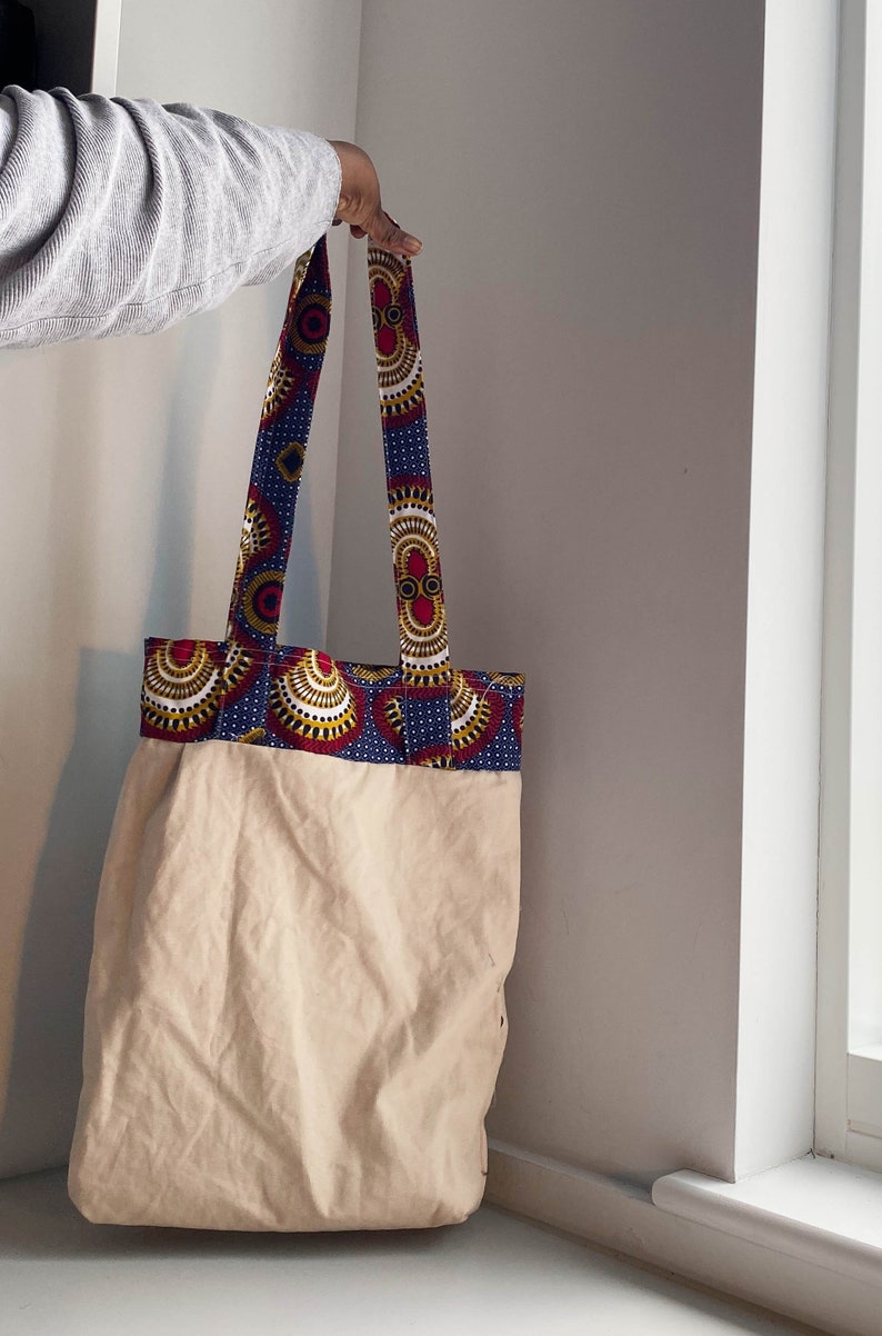 Pode incluir: Uma bolsa tote de lona bege com uma borda de tecido estampado em azul, vermelho e dourado. A bolsa tem duas al&ccedil;as longas.