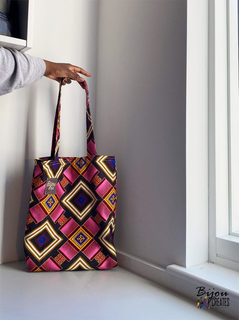 Pode incluir: Uma bolsa tote rosa, amarela, azul e branca com detalhes pretos. A bolsa tem uma etiqueta de tecido preto com o texto "Bijou" em branco.