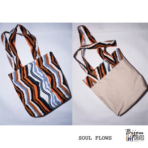 Pode incluir: Uma bolsa tote revers&iacute;vel com um padr&atilde;o ondulado preto, laranja e azul de um lado e uma tela bege lisa do outro. A bolsa tem duas al&ccedil;as longas. O texto "SOUL FLOWS" est&aacute; impresso abaixo da bolsa.
