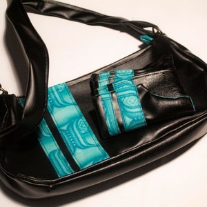 Pode incluir: Uma bolsa de ombro de couro preto com uma aba estampada em turquesa e preto. A bolsa tem um bolso menor que combina na frente.
